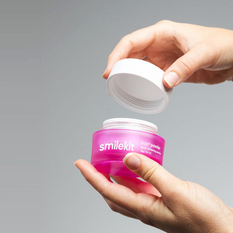 Hands opening a pink Smilekit tooth powder jar against a grey background