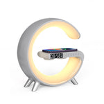 White G-shaped LED lamp glowing warm yellow, with clock display and phone on shelf