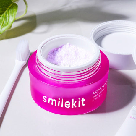 Open pink Smilekit tooth powder jar with white powder, lid and toothbrush nearby